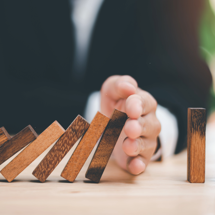 A close-up of a hand in a suit pushing a wooden domino block on a light wooden table, with several dominoes falling in a row.