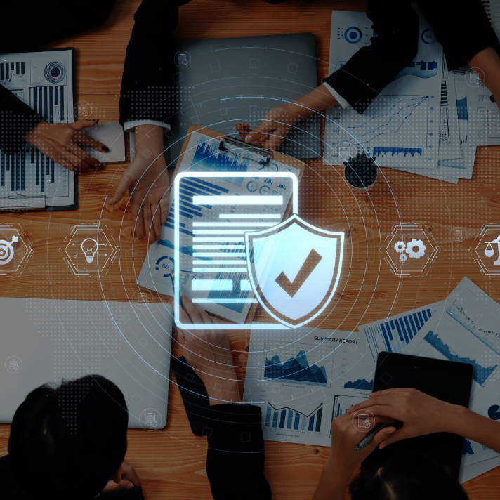 A top-down view of a business meeting around a wooden table, with hands holding laptops, tablets, and printed reports. A glowing digital graphic overlay in the center shows a document icon with a shield and checkmark, surrounded by data charts and icons on a dark blue background.
