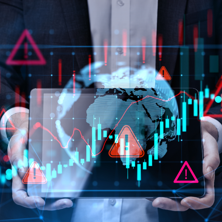A person in a dark suit holds a transparent digital tablet displaying a world map, financial candlestick charts, red warning triangles, and glowing blue data lines over a dark background.