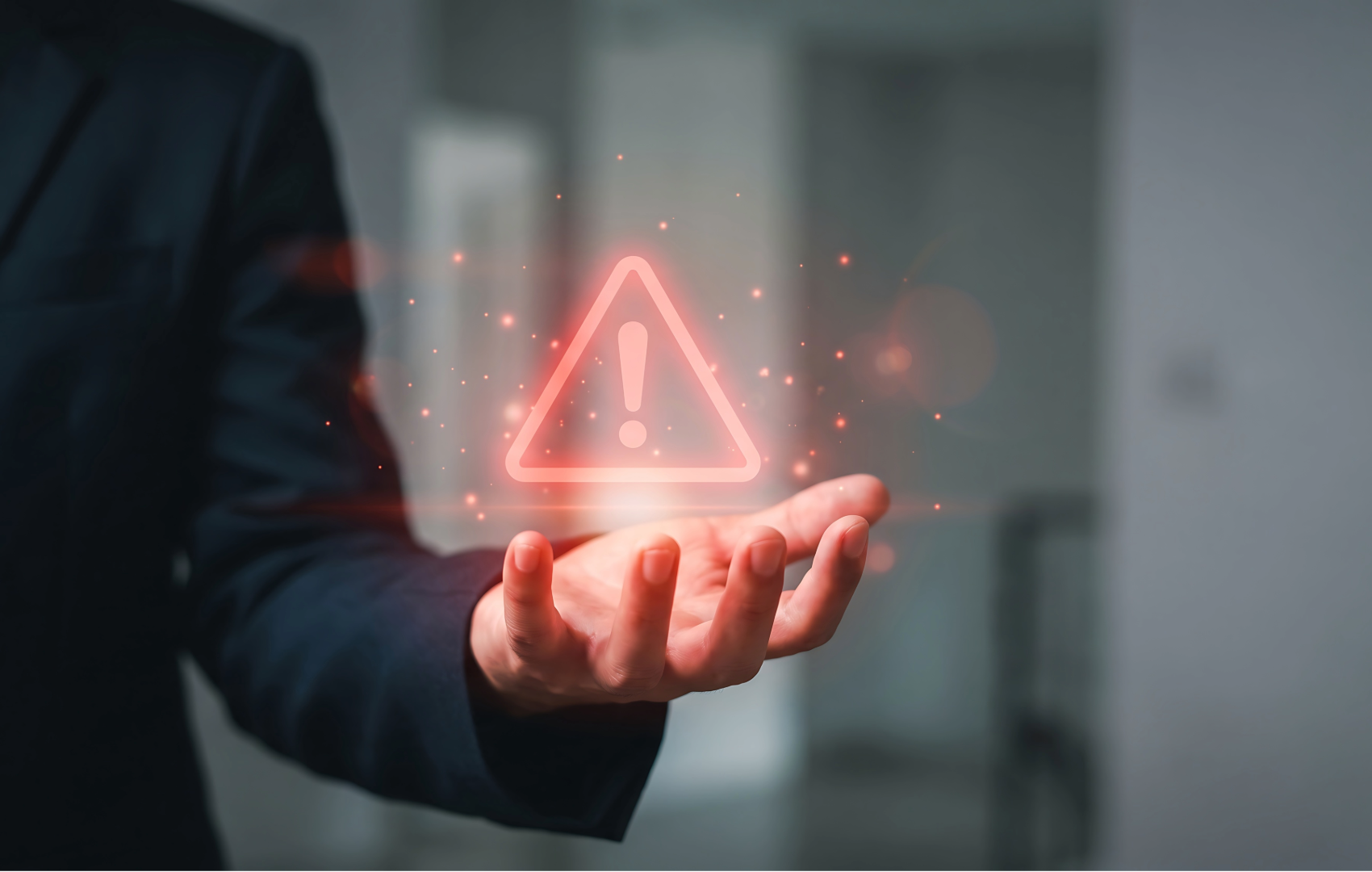 A person in a dark suit extends their hand toward a glowing red triangular warning icon floating in front of a blurred indoor background.