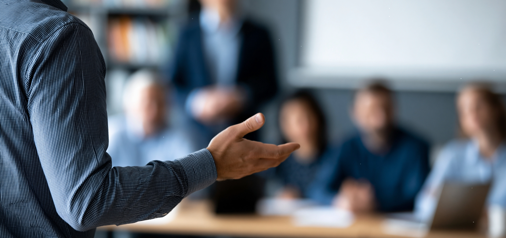 A person in a dark blue striped shirt gestures with their hand during a meeting or presentation, with a blurred office background showing other people and a laptop.