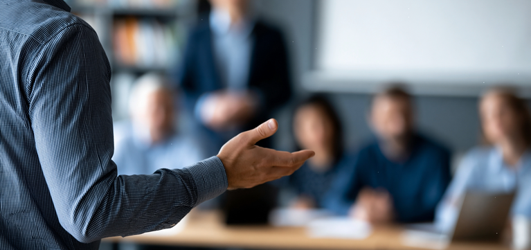 A person in a dark blue striped shirt gestures with their hand during a meeting or presentation, with a blurred office background showing other people and a laptop.